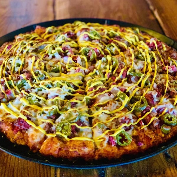 A Parktown Pizza with melted cheese, bacon, jalapeños, and a mustard drizzle on a dark pan.