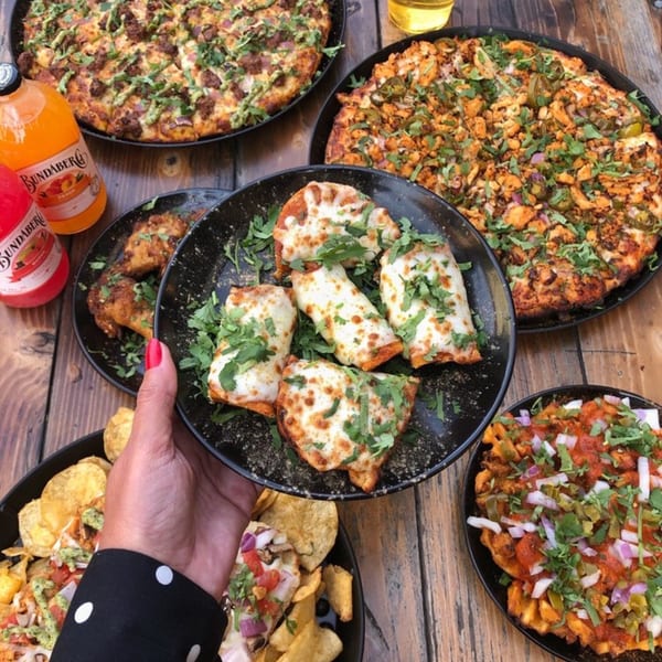 A hand holds a plate of cheesy breadsticks surrounded by pizzas, nachos, and drinks on a wooden table.
