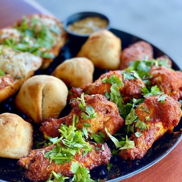 A black platter filled with golden-brown baked dough balls, mini pizzas, and chicken wings garnished with cilantro.