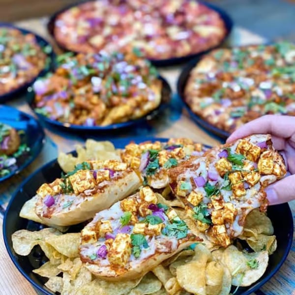 A hand picks up a slice of paneer pizza from a plate of chips, with other pizzas in the background.